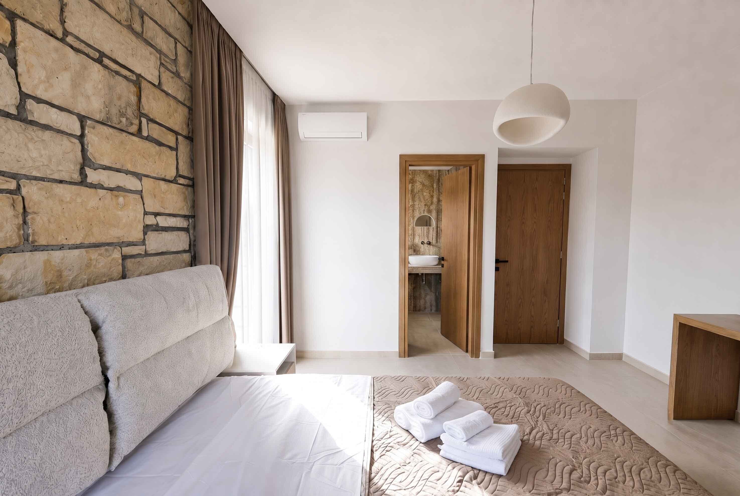 A modern bedroom with a stone wall, a large bed, a bedside table, a hanging light fixture, and a view into the bathroom.