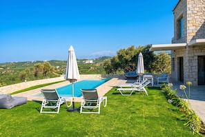 A pool area with lounge chairs and umbrellas, surrounded by a well-maintained lawn and a stone building. - Villa Vision - Holiways Collection (Platanias)