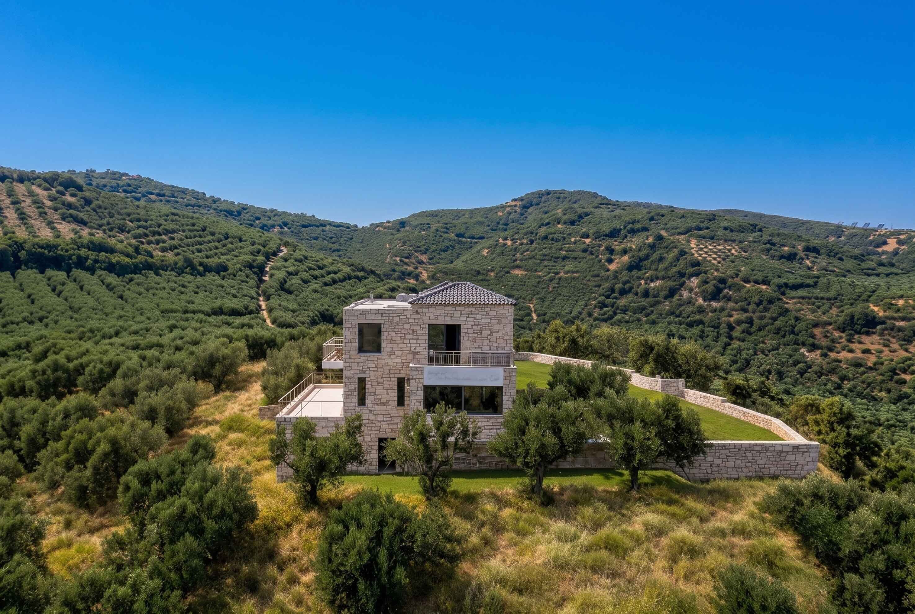 A modern two-story stone house with a balcony, surrounded by a lush landscape and hills.