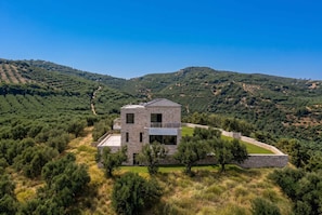 A modern two-story stone house with a balcony, surrounded by a lush landscape and hills. - Villa Vision - Holiways Collection (Platanias)