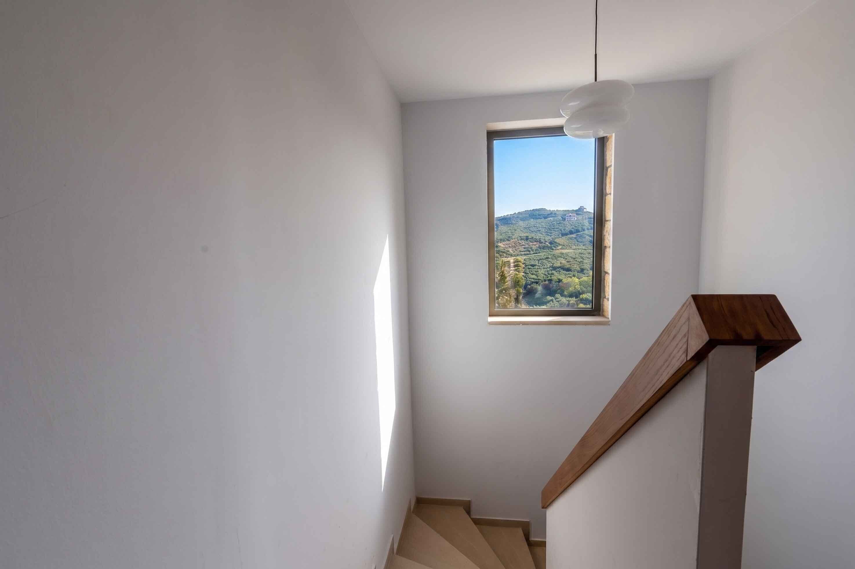 A staircase with a wooden handrail leading to a small window overlooking a hilly landscape.