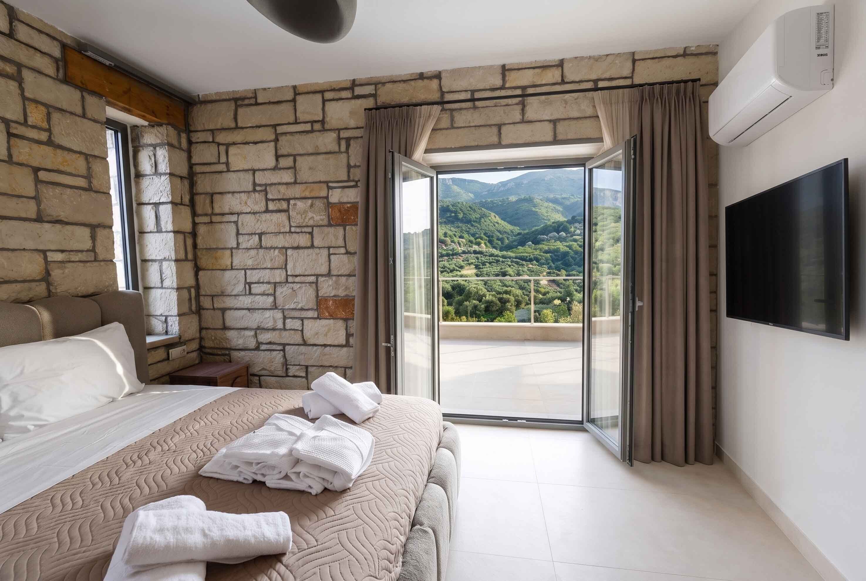 A bedroom with a stone wall, a bed with white linens, a TV on the wall, and a balcony with a mountain view.