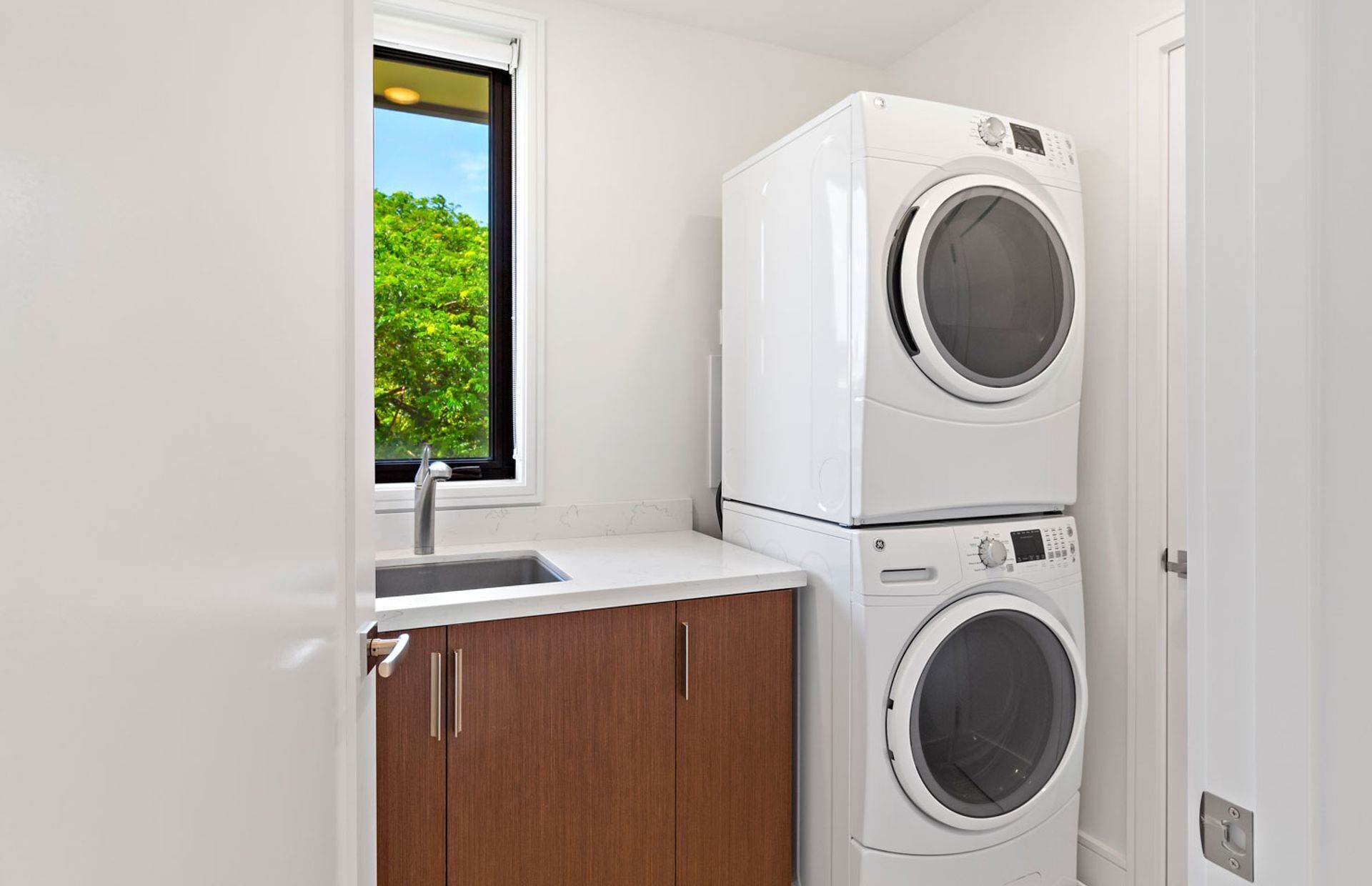 A compact laundry room with a washing machine and dryer stacked on top of each other, a window with a view of greenery, and a sink with a faucet.