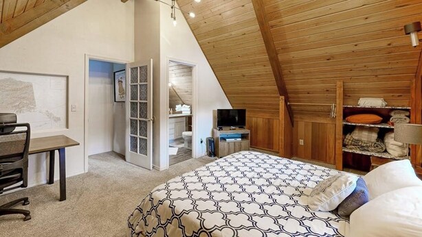 A bedroom with a sloped ceiling, a bed with patterned bedding, a desk with a chair, a television, and a closet with hanging clothes. - Classic Alyeska A-frame From the Resort's Early Days (Girdwood)