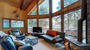 A cozy living room with a fireplace, leather sofas, a flat-screen TV, and large windows offering a view of the forest. - Classic Alyeska A-frame From the Resort's Early Days (Girdwood)