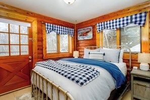 Une chambre dans une cabane en bois, avec un lit en métal, des rideaux à carreaux, une affiche accrochée au mur et une porte en bois.