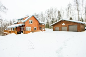 A wooden cabin with a deck and garage, surrounded by snow and trees. - Birch Forest Cabin Retreat W/hot Tub & Anchorage (Chugiak)