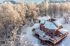 Een houten chalet met terras, omgeven door met sneeuw bedekte bomen en bergen op de achtergrond.