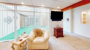 Una habitación con puerta de vidrio corredera que da a un balcón, un sillón de cuero, una pequeña mesa de madera y un televisor montado en la pared.