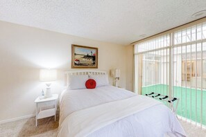 A bedroom with a bed, a nightstand, a lamp, a painting on the wall, and a view through large windows. - Anchorage Flat Between Bootlegger Cove & Downtown (Anchorage)