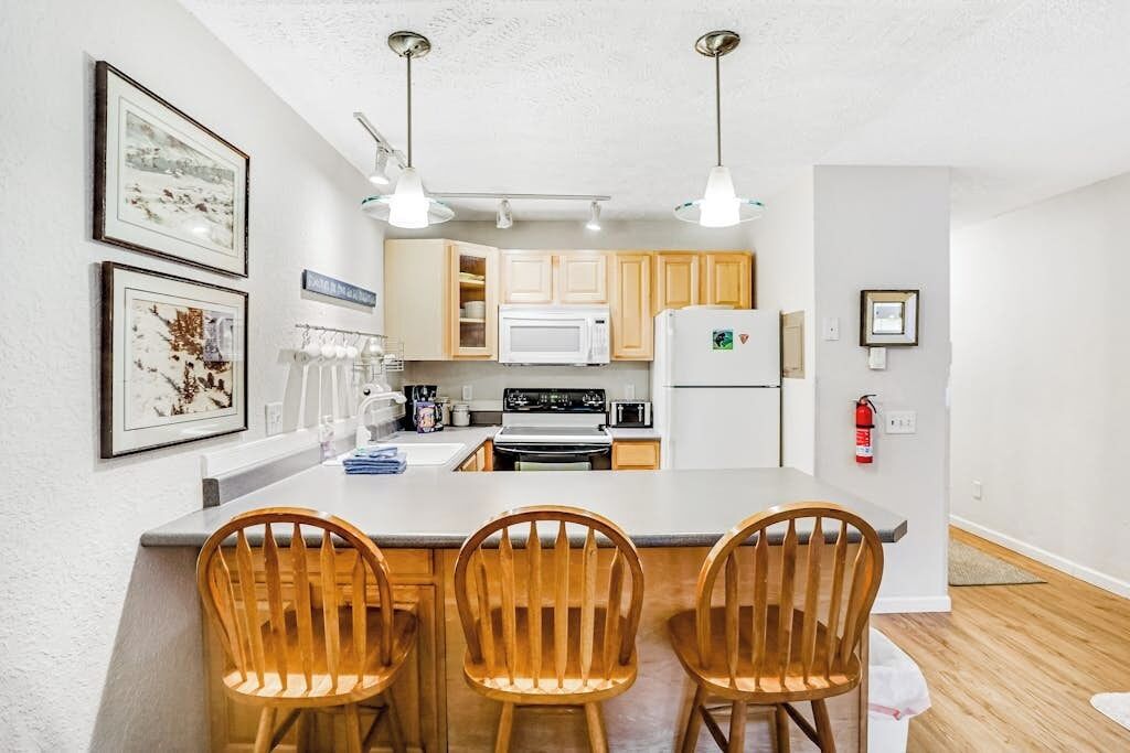 A kitchen with wooden cabinets, a white refrigerator, a microwave, and a stove. There are three wooden chairs at the island.