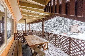 Una terraza de madera con una mesa de picnic y bancos, con vistas a un paisaje nevado con árboles y un edificio.