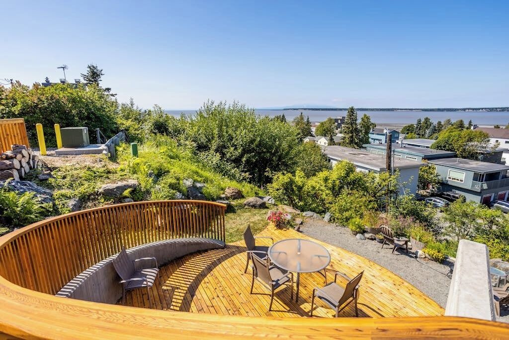 A circular wooden deck with seating and a table, overlooking a body of water and greenery.