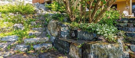 A landscaped area with a stone retaining wall, a variety of plants, and a building in the background.