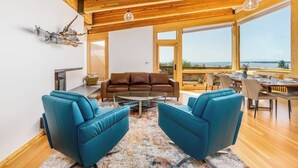 A spacious living area with a wooden ceiling, a brown leather sofa, two blue armchairs, a coffee table, and a dining area with a view of the sea. - Award-winning Anchorage Kitchen w/ Cook Inlet Views (Anchorage)