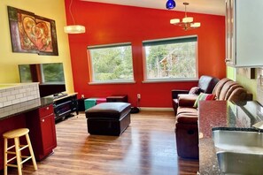 A modern kitchen with a red accent wall, a dining area with a brown leather couch, and a large window providing a view of greenery.