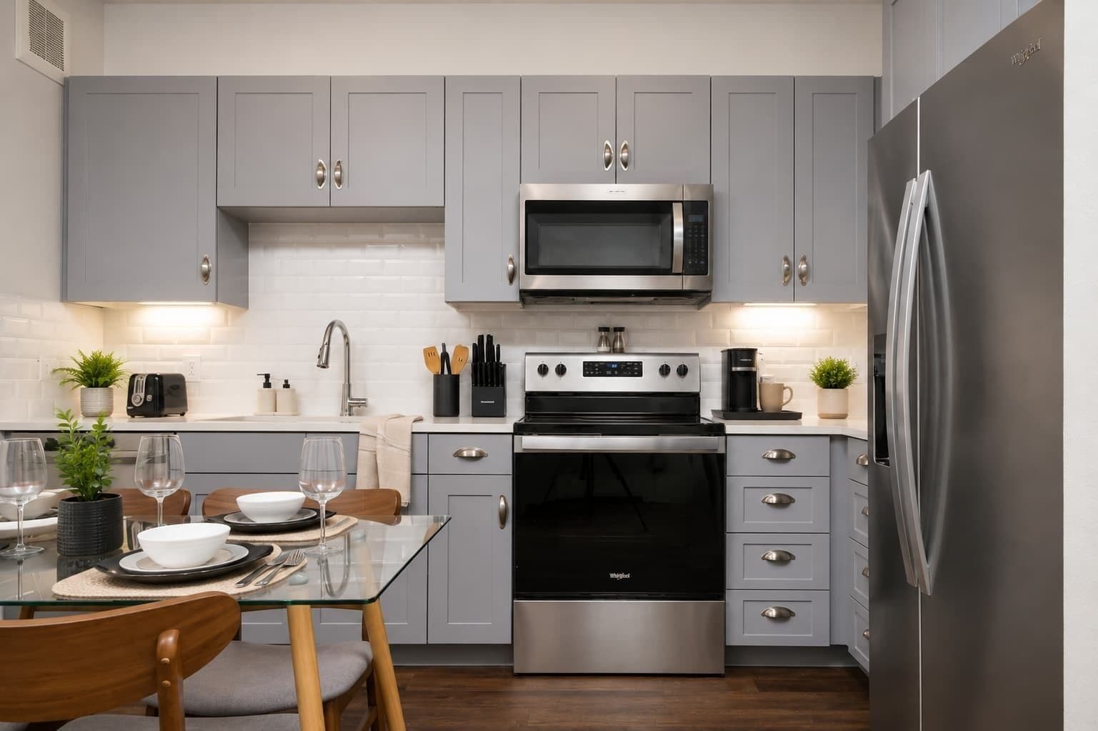 A modern kitchen with grey cabinets, stainless steel appliances, and a glass dining table.