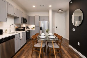 Une cuisine moderne avec des meubles gris, des appareils en acier inoxydable et un coin repas comprenant une table en verre et des chaises en bois.