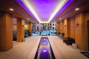 A corridor with a central water feature, wooden walls, and a pool area visible through the glass doors at the end. - Sleek Haven Suite  | Pool • Gym • Parking | Downtown San Antonio (San Antonio)