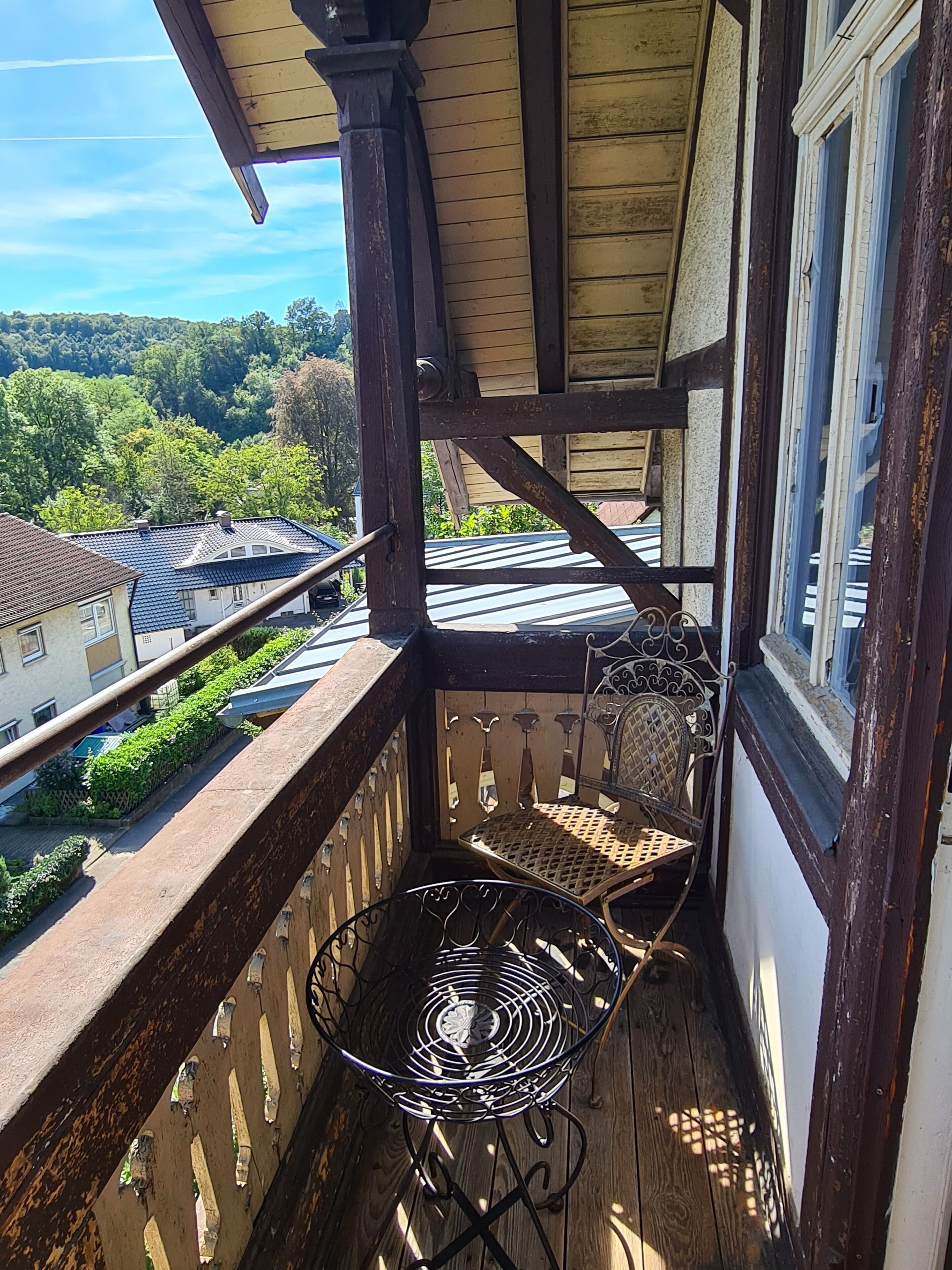 Ein Balkon mit Metallstuhl und Tisch, mit Blick auf eine Wohngegend mit Häusern und Grünflächen.