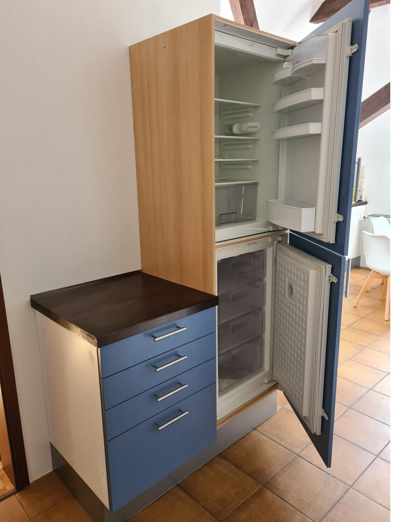 Una cucina compatta con frigorifero integrato, un piano di lavoro in legno e un armadietto blu con cassetti.