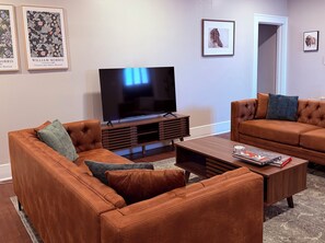 A living room with two brown leather sofas, a wooden coffee table, a flat-screen TV on a wooden stand, and framed artwork on the walls. - Historic 1914 Texas Tudor: Walk to TTU Campus (Lubbock)