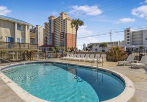 A swimming pool with lounge chairs, a palm tree, and a building in the background. - Gulf View 17: Steps to Beach, Walk to The Hangout, Pool, Pet-Friendly (Gulf Shores)