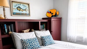 Una cama bien hecha con almohadas estampadas, un cartel de bienvenida enmarcado y un pez decorativo en la pared.