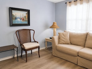 Uma sala de estar com uma cadeira de madeira, um sofá bege, uma mesinha, um abajur e um quadro na parede.