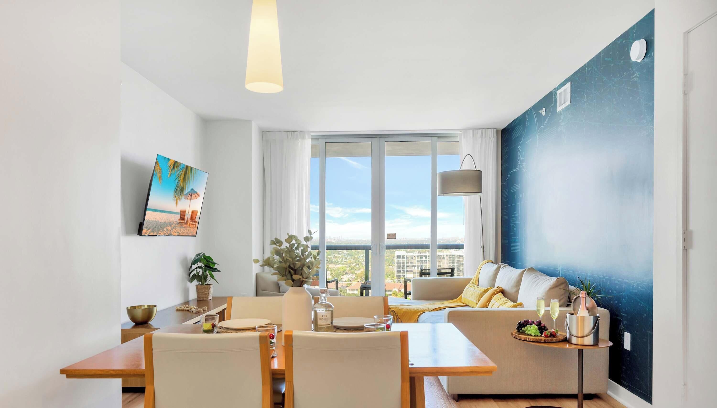 Un salon moderne comprenant un coin repas, une grande fenêtre offrant une vue sur la ville et un mur orné d’un motif abstrait bleu.