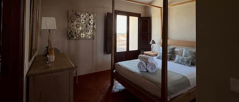 A four-poster bed with rolled towels, a bedside table with a lamp, a decorative wall piece, and a window with a view.