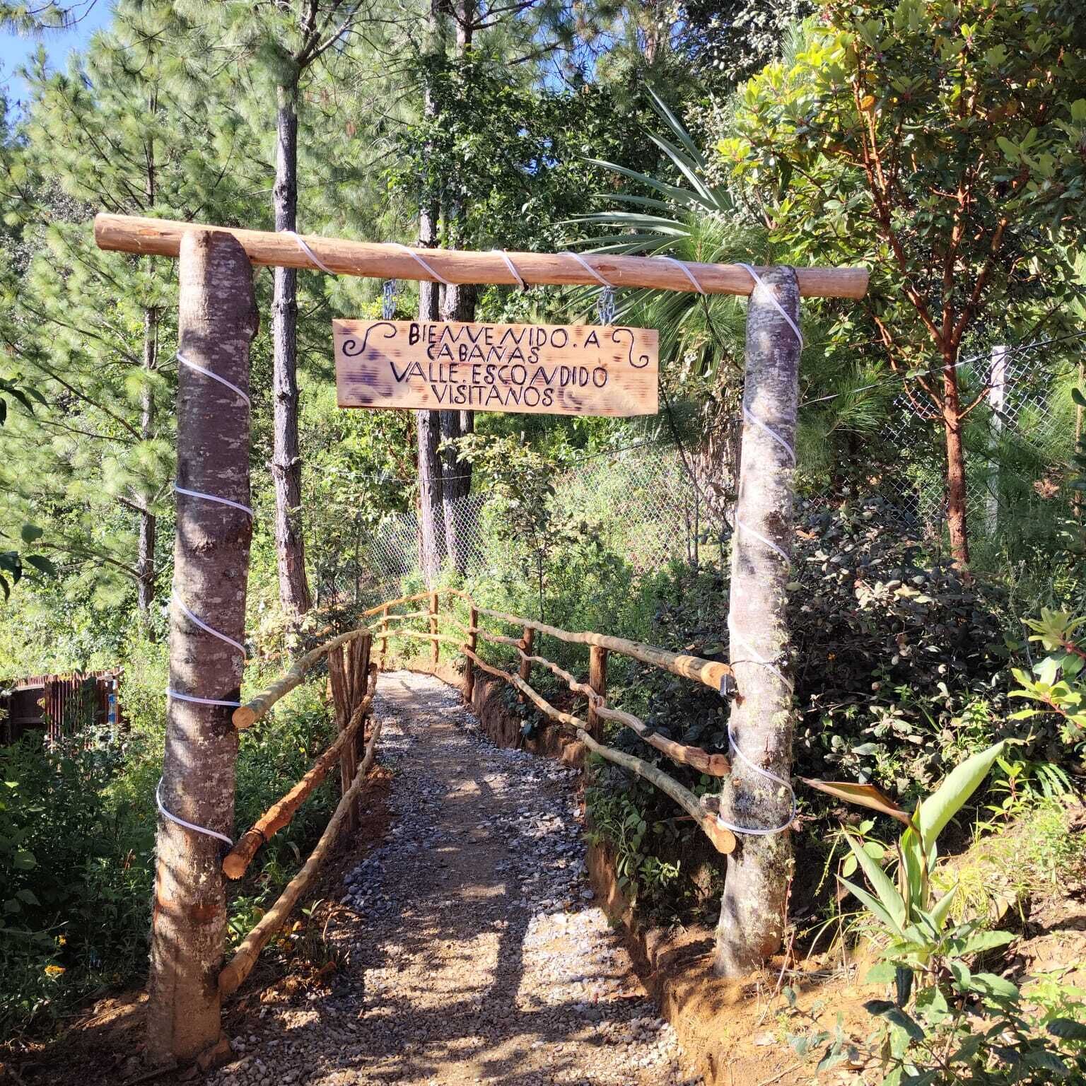 A wooden signpost with a message in Spanish, a rustic wooden bridge, and a forested path.