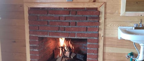 Una chimenea de ladrillo con fuego encendido, una pared de madera y un lavabo blanco con cañerías verdes.