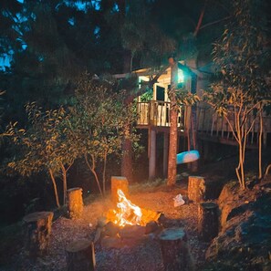 A wooden deck with a fire pit in the center, surrounded by trees and a building with a balcony.