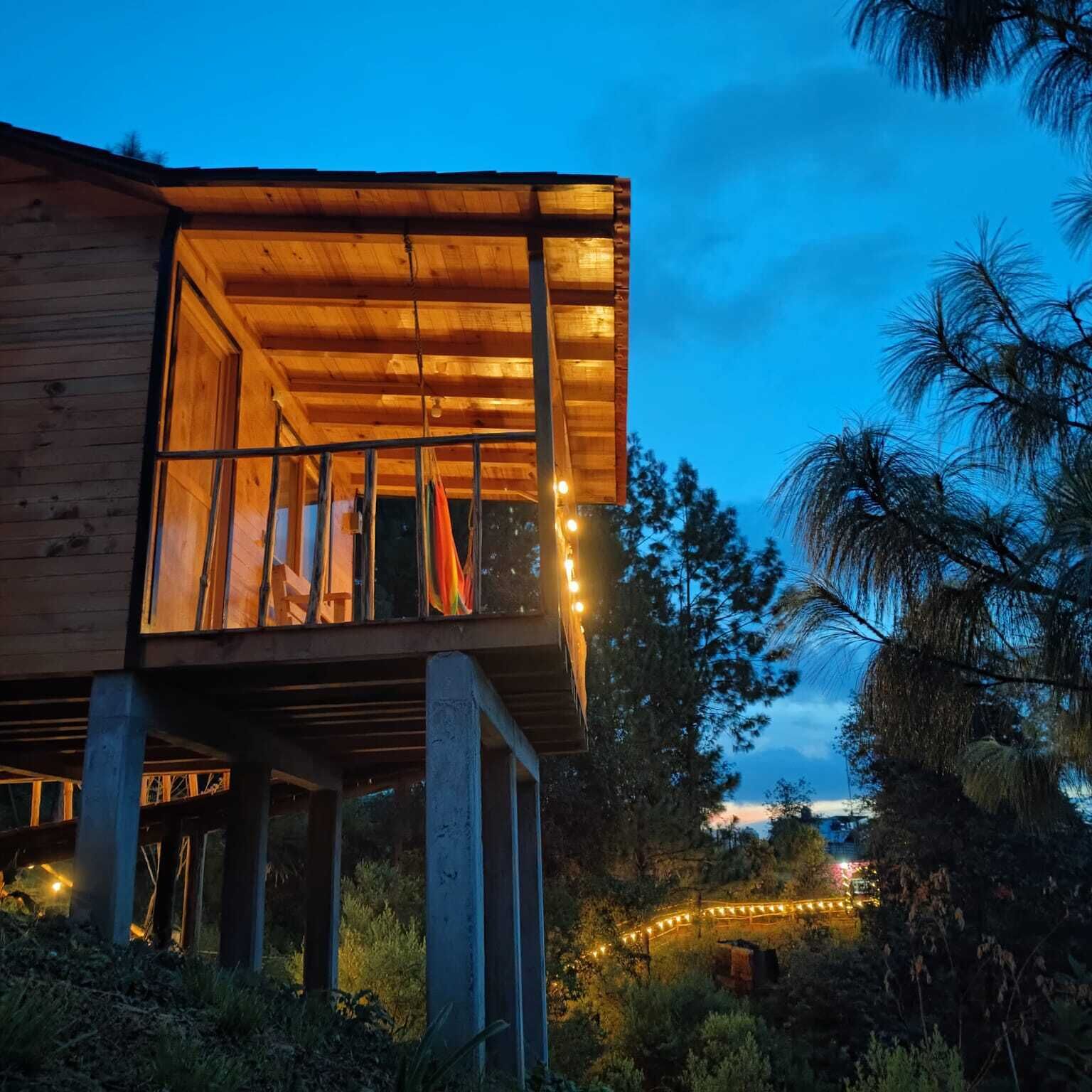 A wooden cabin with a balcony, surrounded by trees and a pathway with lights.
