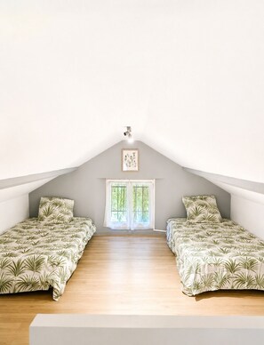 A room with a sloped ceiling, two beds with matching floral bedding, a window with curtains, and a framed picture on the wall. - Petite maisonnette avec accès piscine (Montauban)