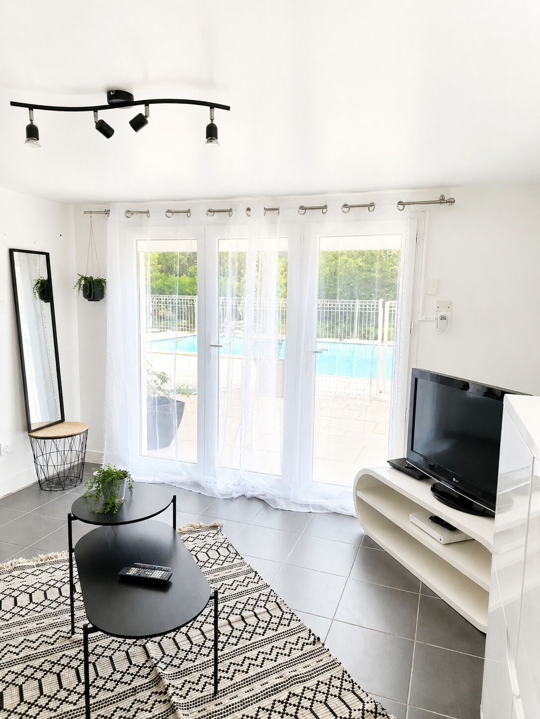 A modern living room with a TV, a dining table, and a large window with a view of a swimming pool.