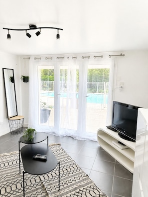 A modern living room with a TV, a dining table, and a large window with a view of a swimming pool. - Petite maisonnette avec accès piscine (Montauban)