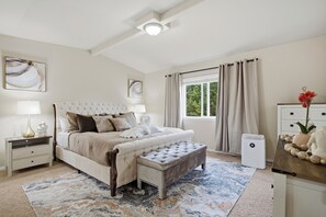 Un dormitorio con una cama grande, mesitas de noche, una banca, una ventana con cortinas y un radiador.