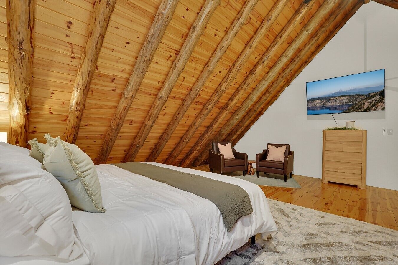 A bedroom with a sloped ceiling, a bed, two armchairs, a television, and a wooden cabinet.