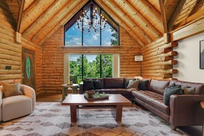 A log cabin interior with a large window, a chandelier, a leather sofa, and a wooden coffee table.