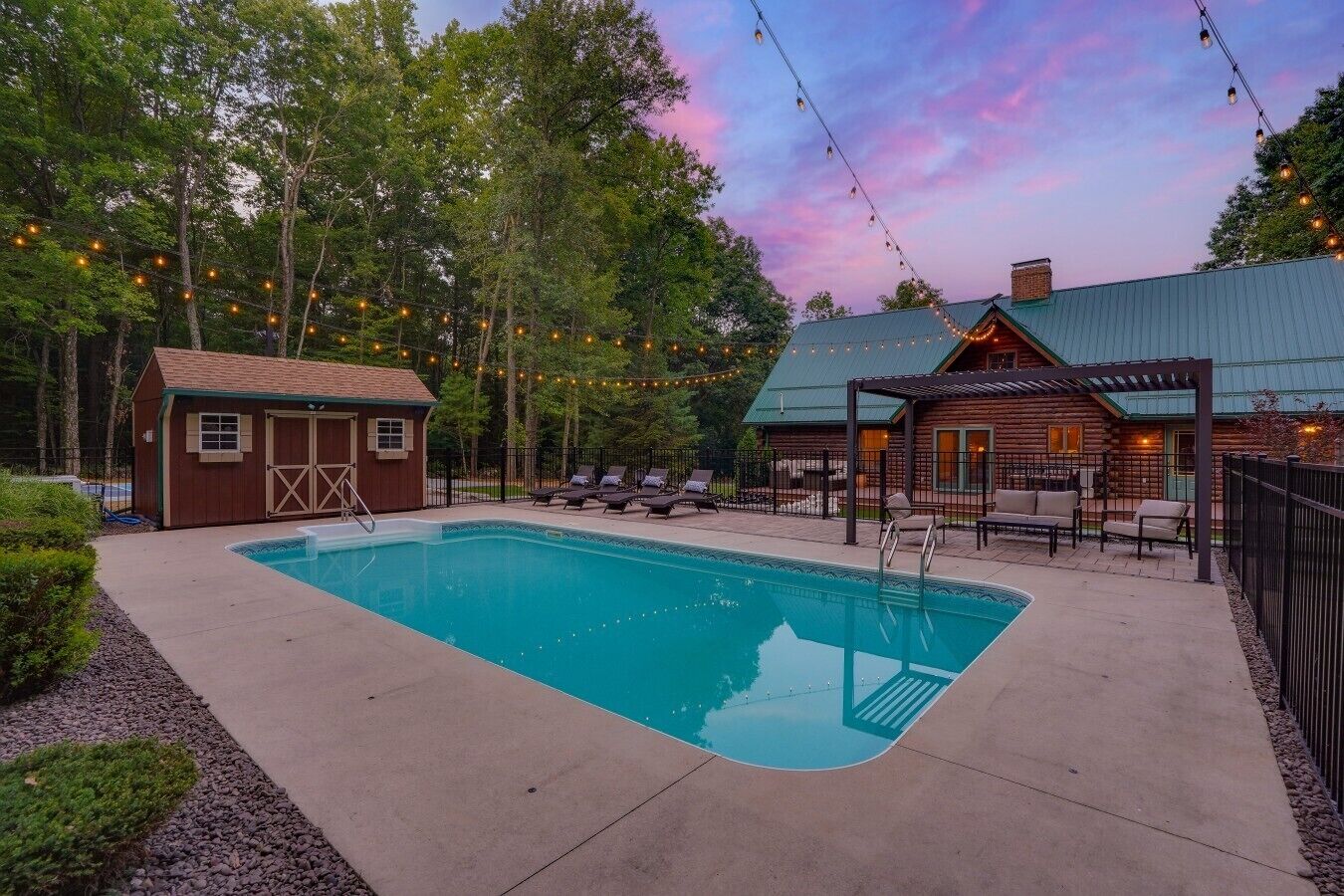 A swimming pool with lounge chairs and a wooden cabin, surrounded by trees and string lights.