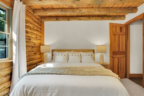Un dormitorio con pared de troncos de madera, una cama con ropa de cama blanca, dos lámparas de noche y una puerta de madera.