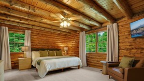 A log cabin bedroom with a bed, bedside tables, a sofa, a ceiling fan, and a window with curtains. - Pine Mountain Lodge - Pool - Pickleball (Fayetteville)
