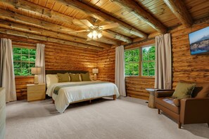 Un dormitorio tipo cabaña de madera con una cama, mesitas de noche, un sofá, un ventilador de techo y una ventana con cortinas.