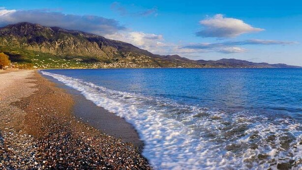 Una playa con piedras, agua tranquila y montañas al fondo.