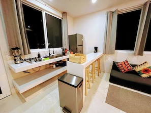 A compact kitchenette with a gas stove, sink, and a compact refrigerator. There is a small bar stool and a black trash bin. - Mangal Suites Vasa (Brisas de Zicatela)