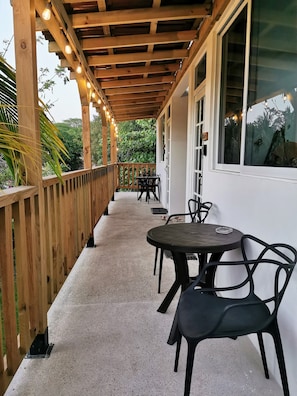 A covered outdoor area with a wooden deck, a seating arrangement with tables and chairs, and a window with a view of greenery. - Mangal Suites Vasa (Brisas de Zicatela)