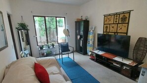 Una sala de estar con sofá, televisor, zona de comedor con mesa y sillas, y un ventanal grande.