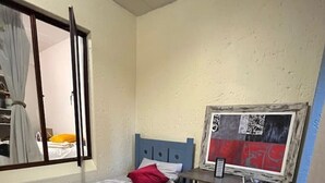 Una habitación pequeña y funcional con una cama individual, un escritorio con computadora y una ventana con cortinas.